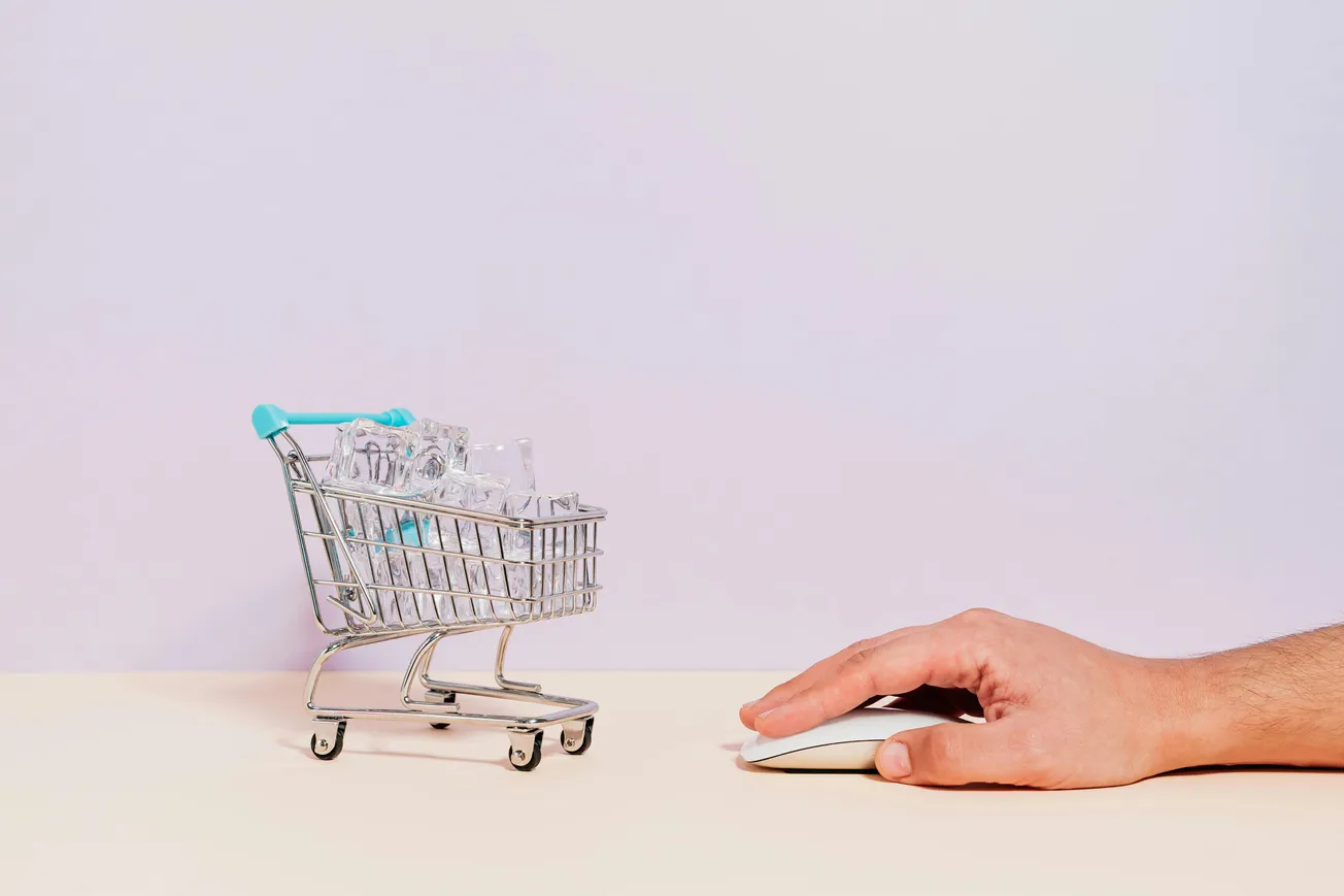 A small shopping cart filled with boxes is on a beige surface. Beside it, a hand uses a computer mouse, symbolizing online shopping. The tone is modern and digital.