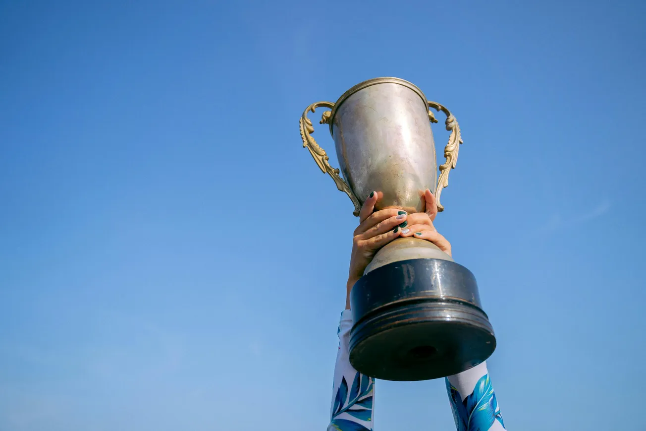 Hands triumphantly raise a large silver trophy against a clear blue sky. The scene conveys victory and celebration, with a tone of achievement.