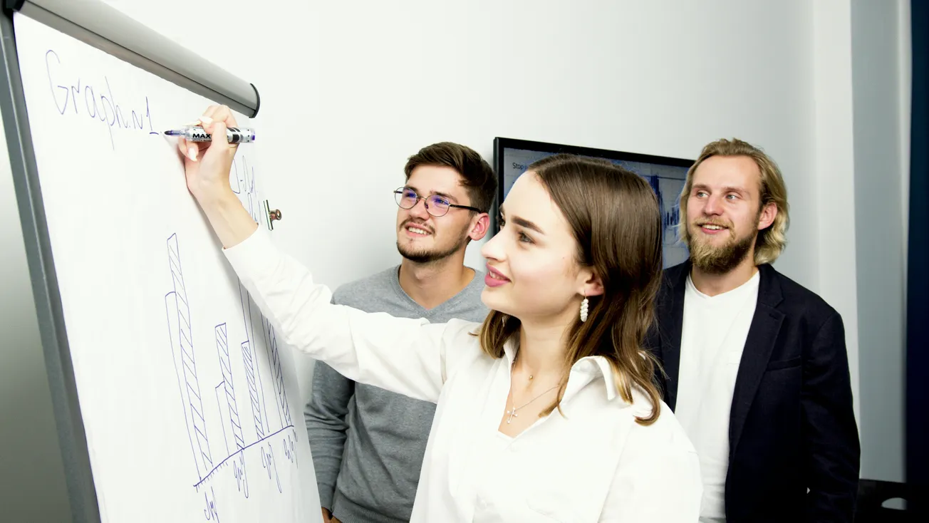 A woman in a white blouse writes on a flip chart with bar graphs as two men observe. They appear engaged and focused in a bright office setting.