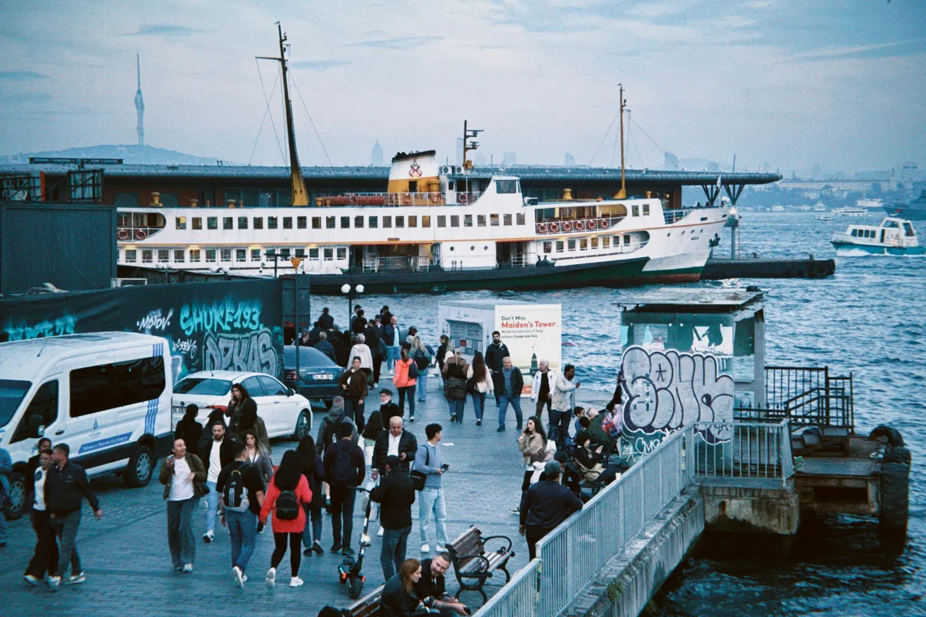 A bustling dock scene with people walking and chatting near a graffiti-covered wall. A large ferry is docked against a backdrop of water and a distant cityscape.