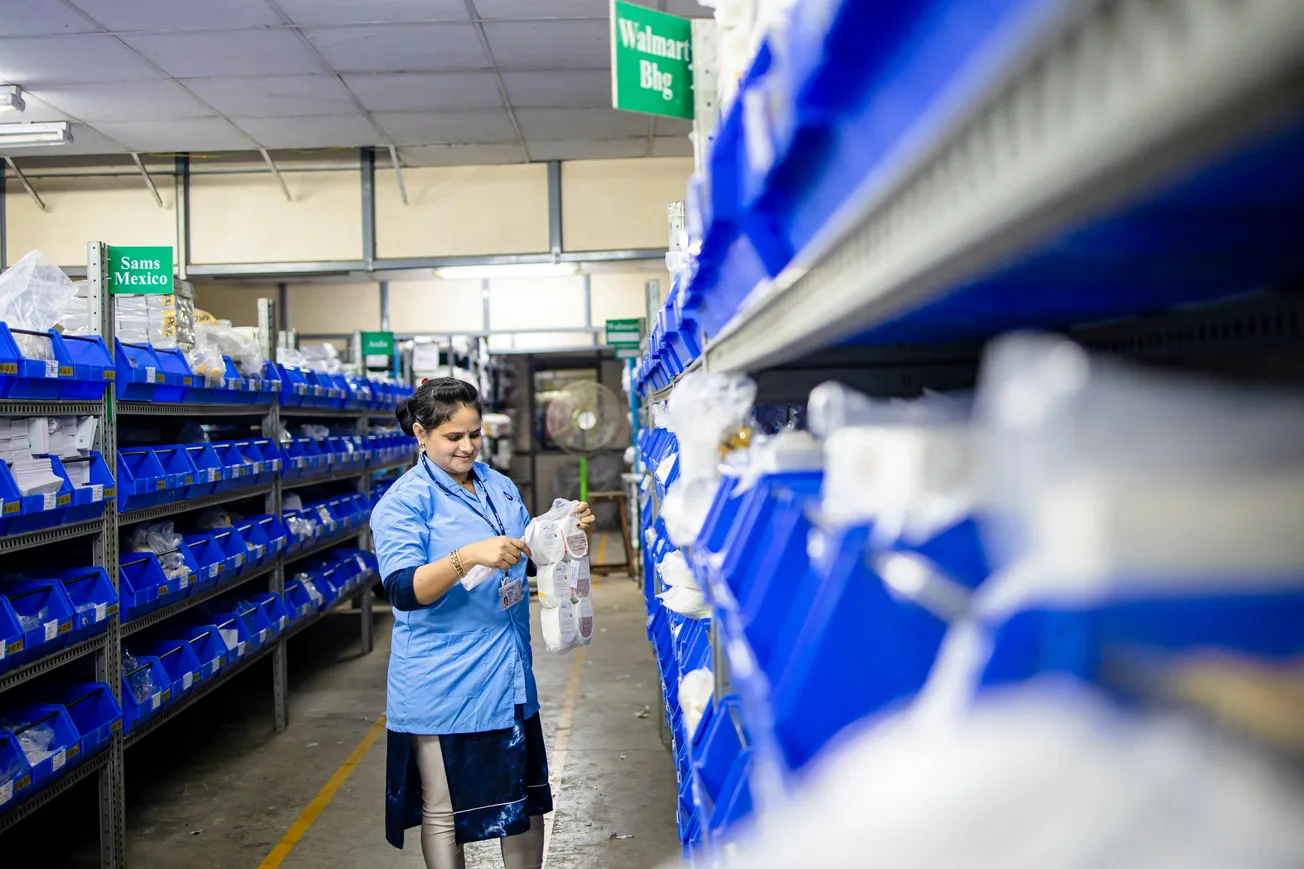 A woman in a blue uniform inspects items in a warehouse aisle, surrounded by organized blue bins on shelves. The scene conveys focus and efficiency.