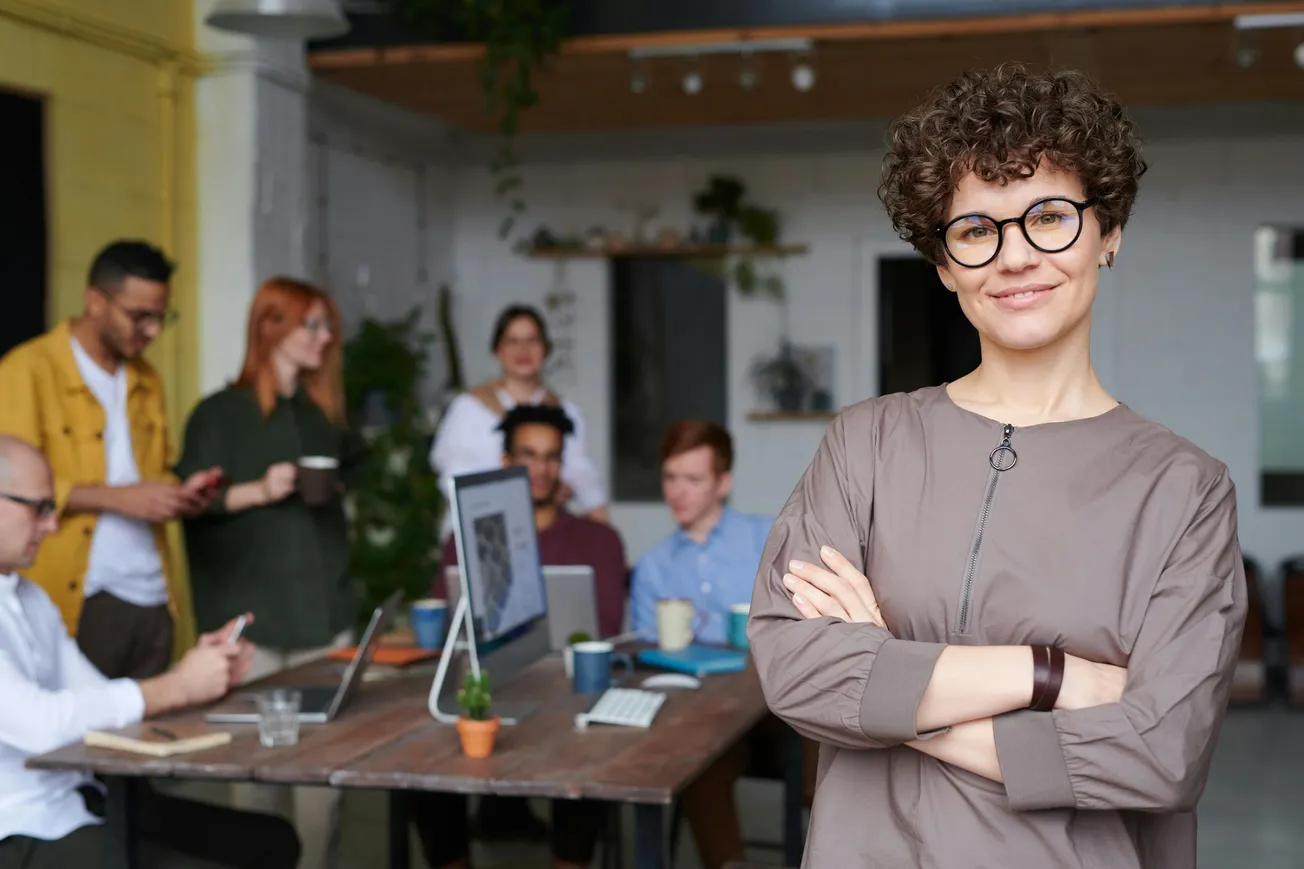 A confident person with curly hair and glasses stands smiling in the foreground. In the background, a diverse group engages in a team meeting.