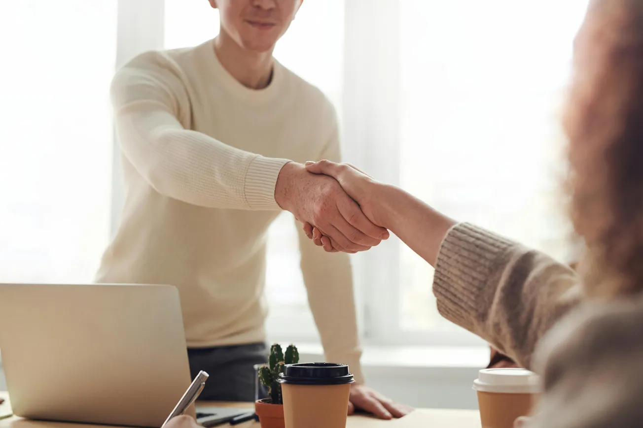 A person in a cream sweater and another in a beige sweater share a handshake in a bright office. A laptop, coffee cups, and a notepad are on the table.