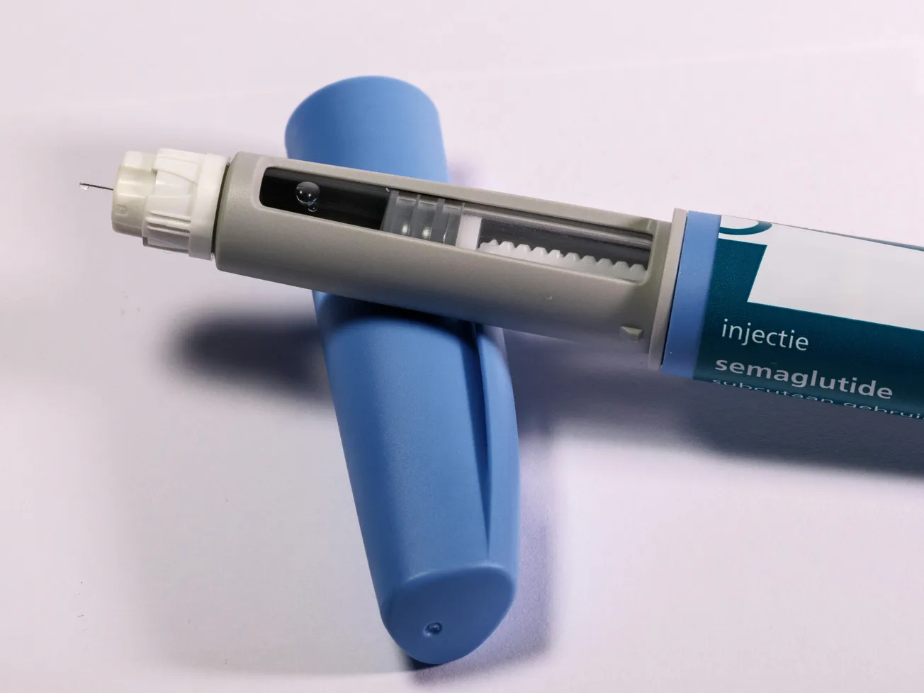 A close-up of a semaglutide injector pen with its blue cap removed, revealing a small needle. The pen's body shows dosage markings.