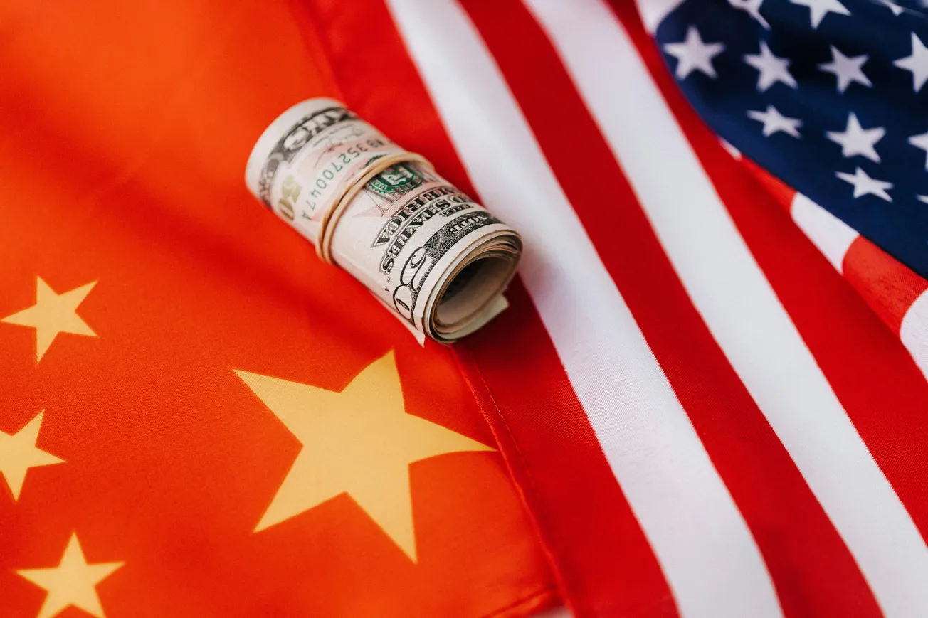 Rolled U.S. dollar bills sit between partially visible Chinese and American flags, symbolizing international economic relations.