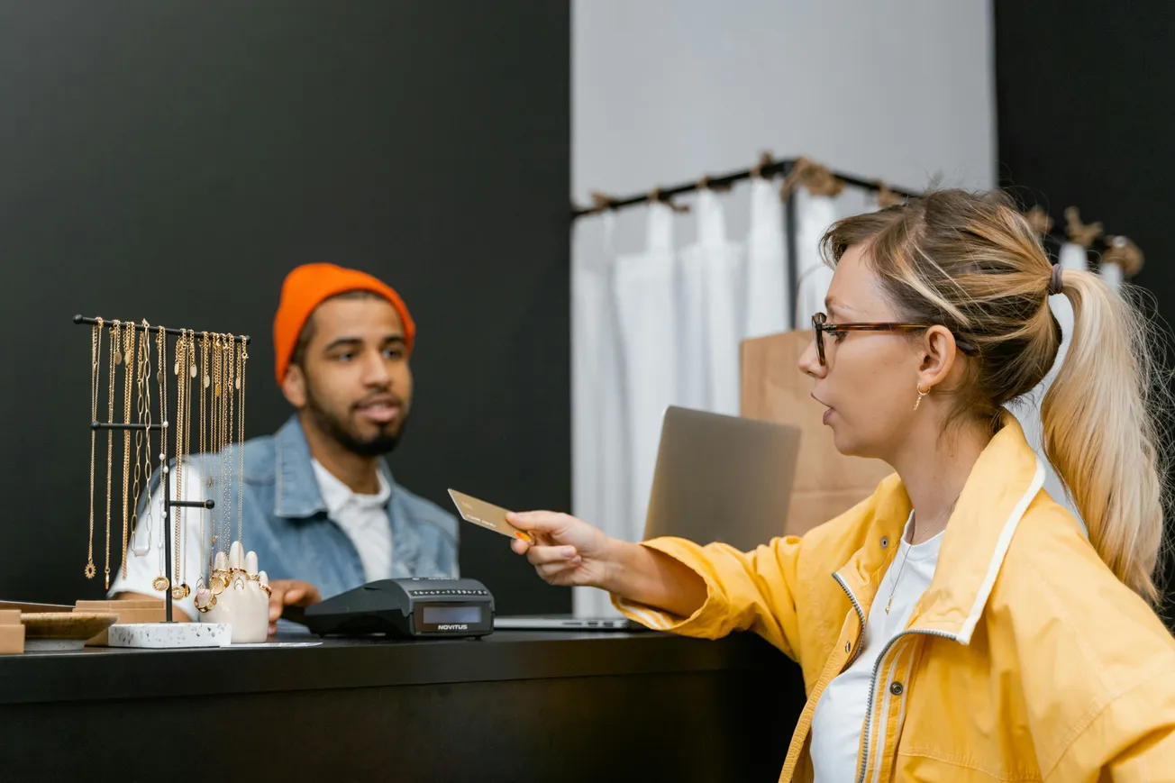 A woman in a yellow jacket hands a credit card to a bearded cashier in an orange beanie at a jewelry counter. The scene conveys a casual, focused interaction.