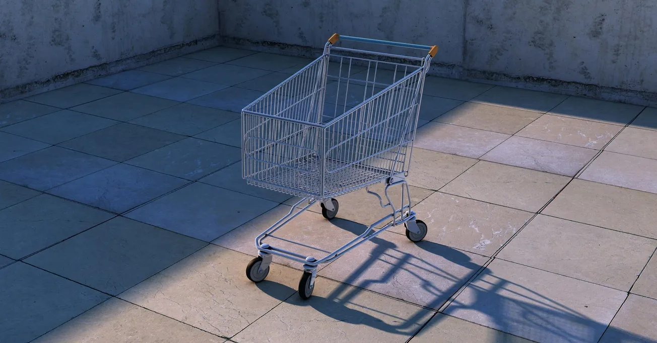 A solitary shopping cart stands on a sunlit, tiled surface near a concrete wall. The cart casts a clear shadow, conveying a sense of emptiness and isolation.