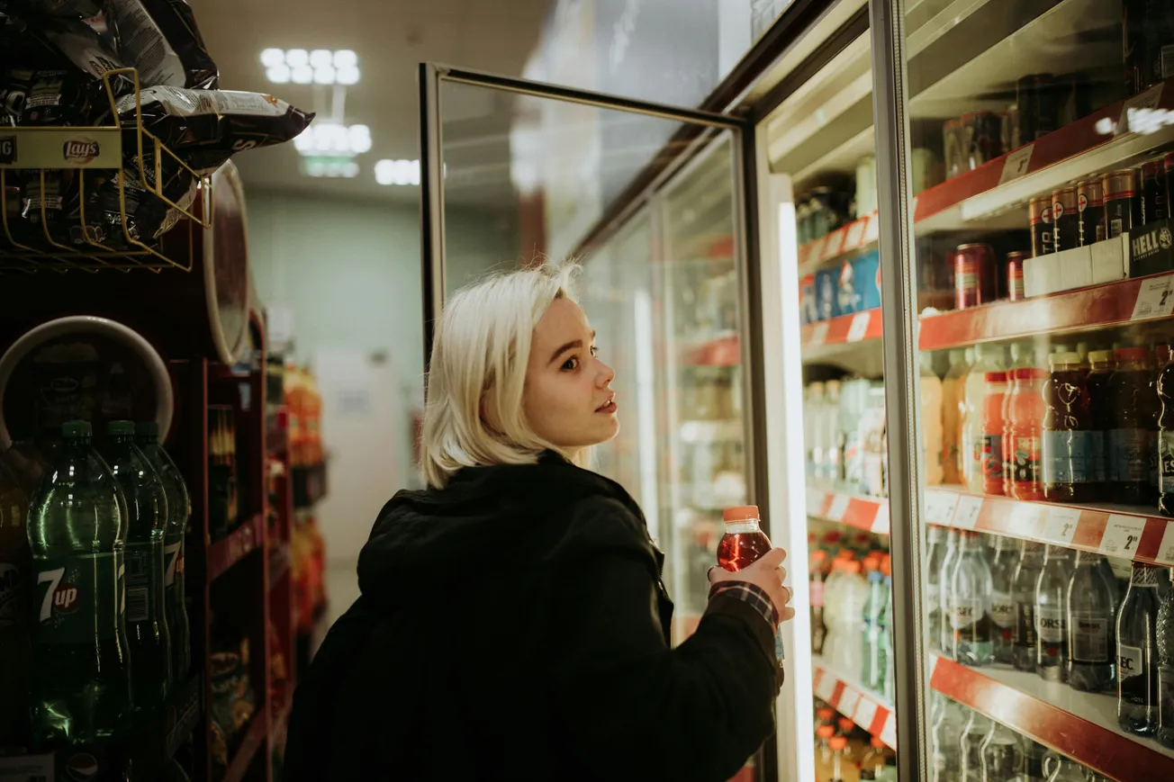 A person with short blonde hair is holding a drink and looking at a well-lit beverage fridge in a grocery aisle. The setting seems casual and everyday.