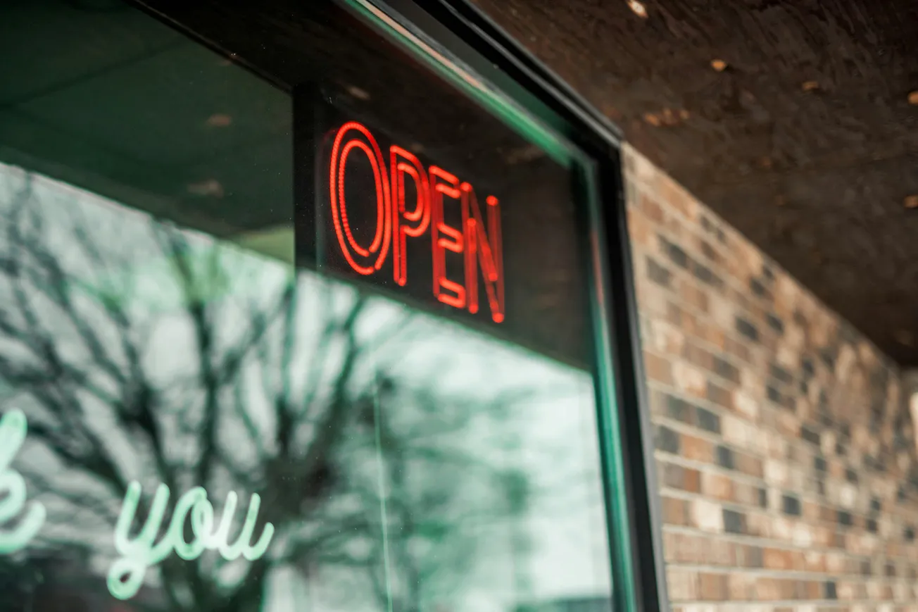 Red neon "OPEN" sign in a window reflects tree branches. The adjacent brick wall adds texture, conveying a welcoming, urban atmosphere.