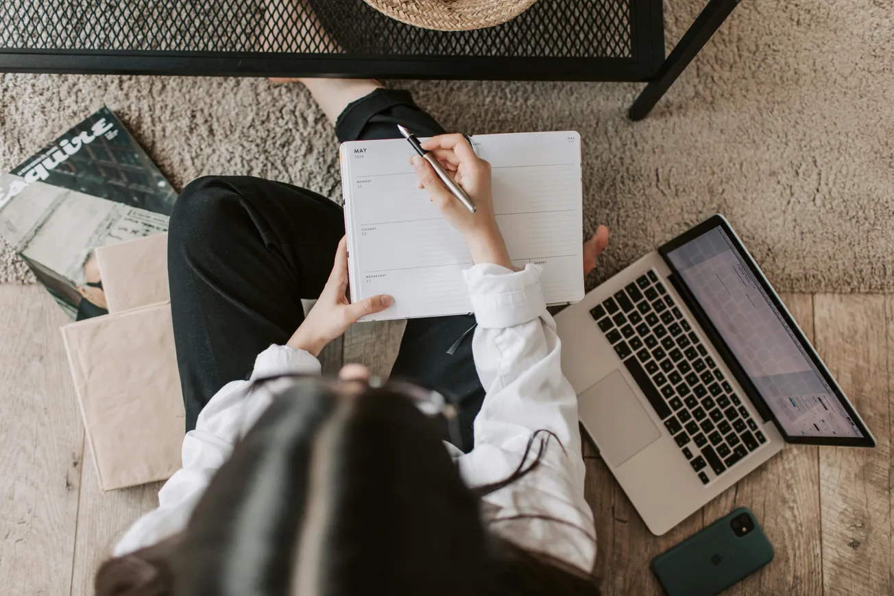 Person seated on the floor, using a laptop and writing in a planner. Nearby, a phone and magazines are placed. The scene is calm and organized.