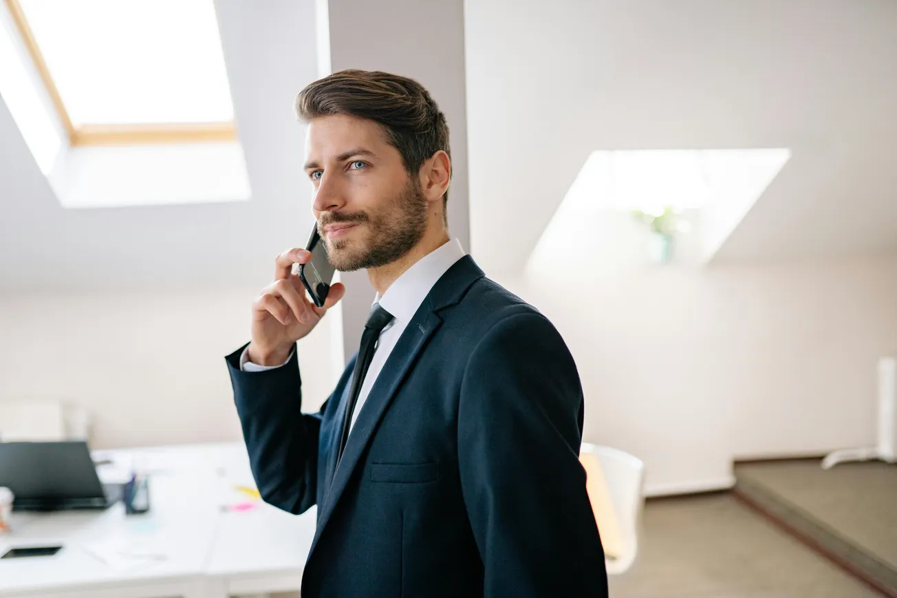A man in a suit smiles while talking on a phone in a bright office with skylights. The scene conveys a professional and upbeat atmosphere.